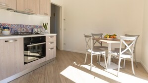 Une cuisine moderne équipée d’une cuisinière à gaz, d’un four et de meubles blancs. On y trouve une table à manger avec des chaises blanches et un panier de fruits.