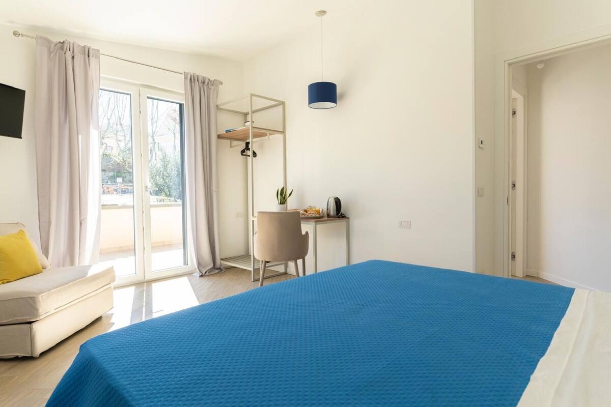 Une chambre avec un grand lit, un bureau avec une chaise, une plante en pot et une vue sur l’extérieur.
