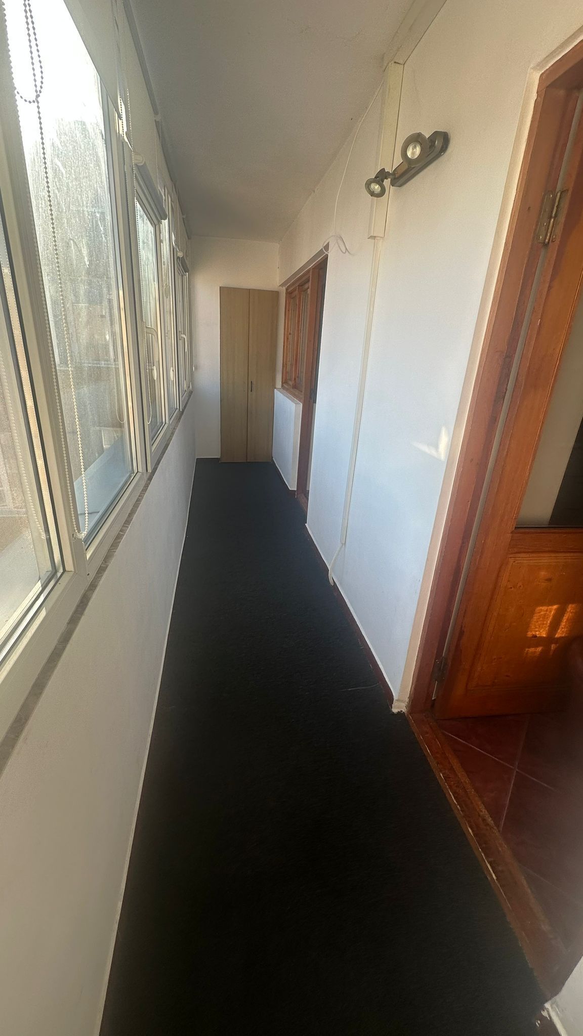 Un couloir étroit, doté d’un tapis noir, de portes en bois et d’une fenêtre avec des stores.