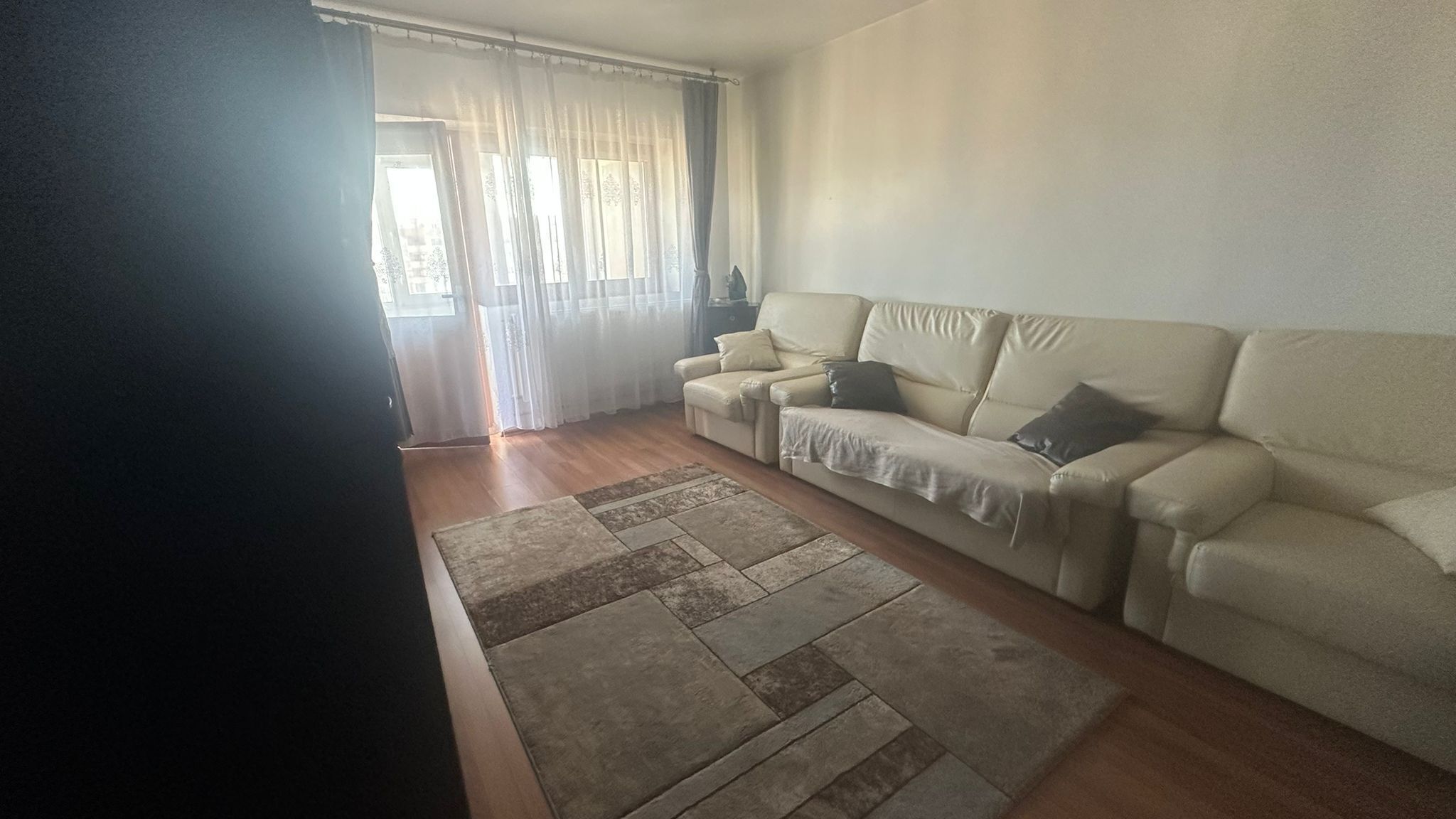 En stue med en beige sofa, et mønstret tæppe og et vindue med lette gardiner.