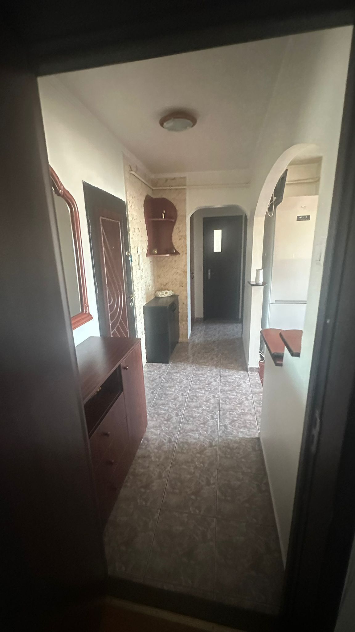 A hallway with a tiled floor, a wooden cabinet, a small trash bin, and a door at the end.