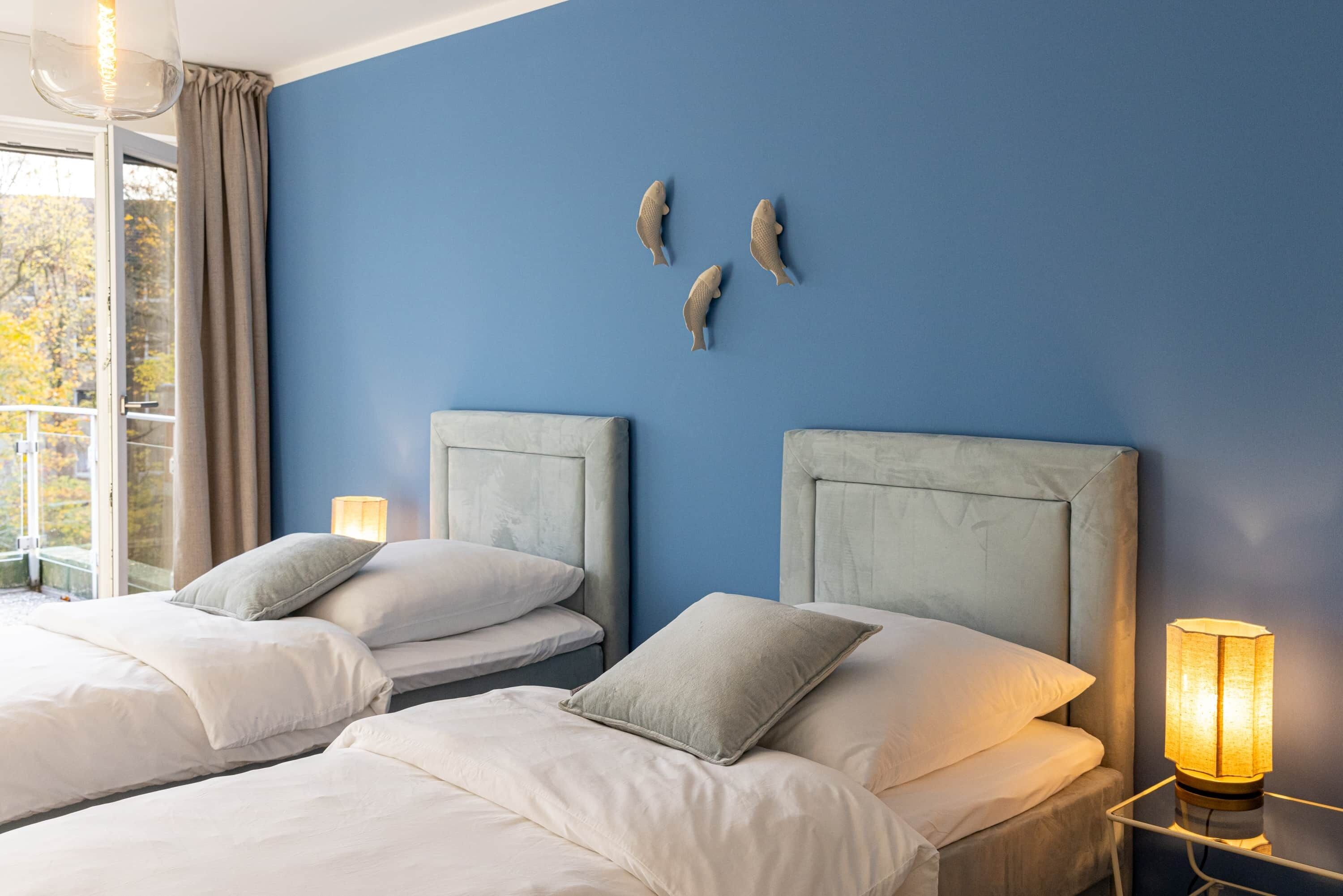 A hotel room with two beds, a large window with curtains, a nightstand with a lamp, and wall art.