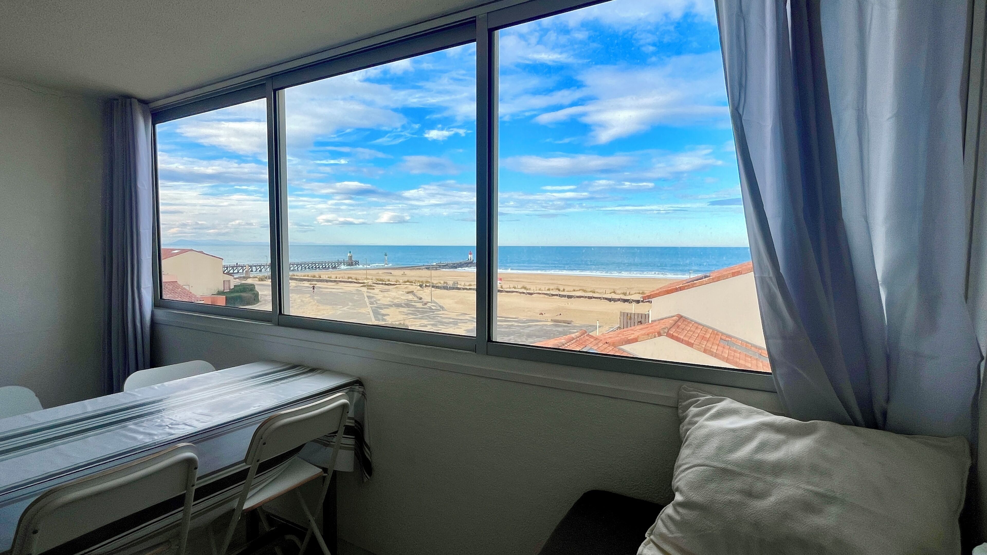 Une chambre dotée d’une grande fenêtre donnant sur la plage et une jetée. On y trouve un lit avec un oreiller blanc et une petite table avec une chaise.