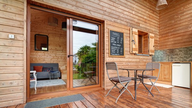 A wooden cabin interior with a sliding glass door leading to a patio, a dining area with metal chairs, a small kitchen with a sink, and a chalkboard menu. - Chalet in Périgord Vert (Sainte-Croix de Mareuil)