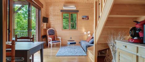 A wooden interior with a dining table, chairs, a sofa, a TV, and a window with a view of greenery.