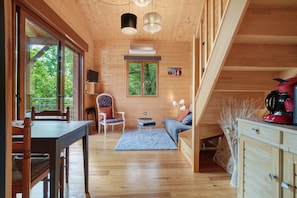 A wooden interior with a dining table, chairs, a sofa, a TV, and a window with a view of greenery. - Chalet in Périgord Vert (Sainte-Croix de Mareuil)