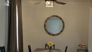 Een eetruimte met een tafel, stoelen, een plafondventilator, een spiegel en een bijzettafeltje met bloemen.