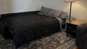 Een kleine, verzorgde slaapkamer met een bed, een nachtkastje, een lamp en een vloerkleed.