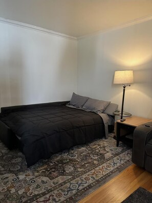 Ein kleines, ordentlich eingerichtetes Schlafzimmer mit Bett, Nachttisch, Lampe und Teppich.