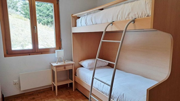 Kerrossängyllä varustettu huone, jossa on puinen tikapuu, pieni yöpöytä lampun kanssa ja ikkuna, josta avautuu näkymä vehreään maisemaan.
