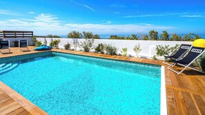 Piscina azul límpida com deck de madeira, espreguiçadeiras e parede branca com vegetação.