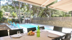 A dining table set for four with plates, cutlery, and a bottle of water. A pool is visible in the background, surrounded by trees and a stone wall. - Piscine/ Proche mer et village / Carry Le Rouet (Carry-le-Rouet)