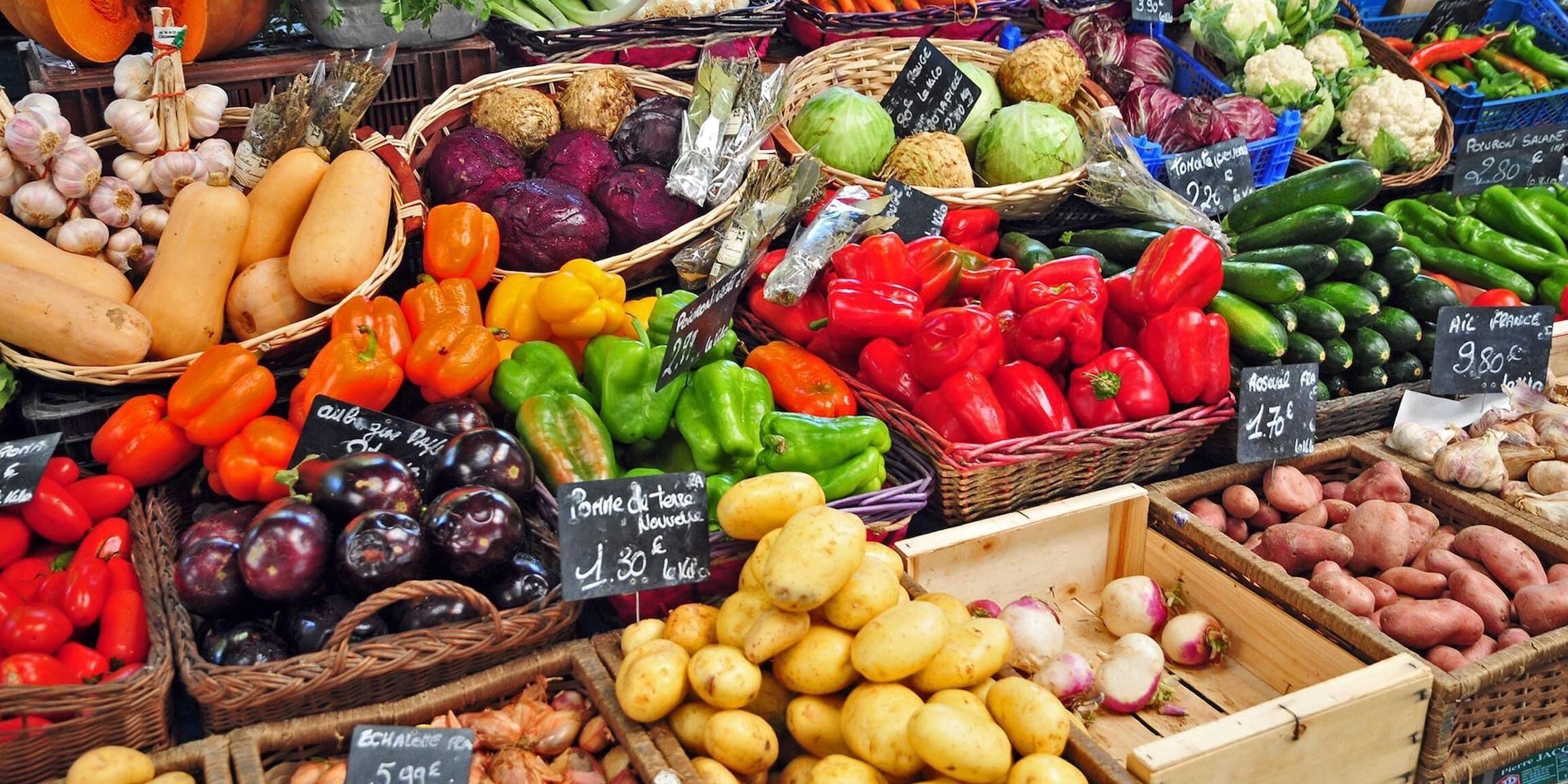 Un puesto de mercado con diversos vegetales y frutas, incluyendo pimientos, berenjenas y papas, expuestos en cestas y cajas.