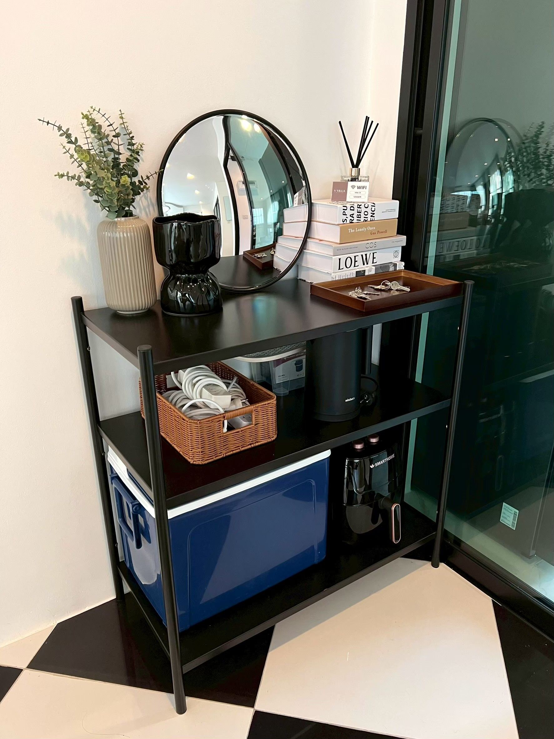 A black metal console table with a mirror, a vase with green foliage, a stack of books, and a tray with keys.