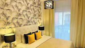 Quarto de hotel com cabeceira estofada, cama grande com almofadas amarelas e pretas, duas luminárias de apoio, papel de parede estampado e vista para o exterior através de uma janela com cortinas.