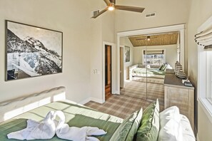 Ein Schlafzimmer mit einem Bett, einem Deckenventilator, einem großen gerahmten Bild und Blick in einen angrenzenden Raum mit einer Couch und einem Fenster.