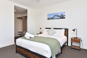 A hotel room with a bed, a nightstand, a lamp, a painting of a bridge, and a closet. - Oceanview Penthouse Caves Beach (Caves Beach)