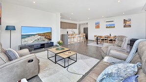 A modern living room with a flat-screen TV, a sofa, a coffee table, and a dining area with chairs and a table. - Oceanview Penthouse Caves Beach (Caves Beach)