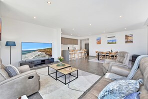 A modern living room with a flat-screen TV, a sofa, a coffee table, and a dining area with chairs and a table. - Oceanview Penthouse Caves Beach (Caves Beach)