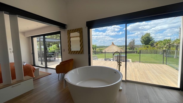 Un baño moderno con una bañera independiente, puertas correderas de vidrio que dan a un patio con mobiliario exterior y vistas a una zona ajardinada con un pabellón.