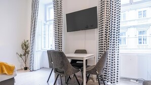 Una habitación moderna con una mesa blanca y dos sillas, un televisor de pantalla plana en la pared y una ventana con cortinas estampadas.