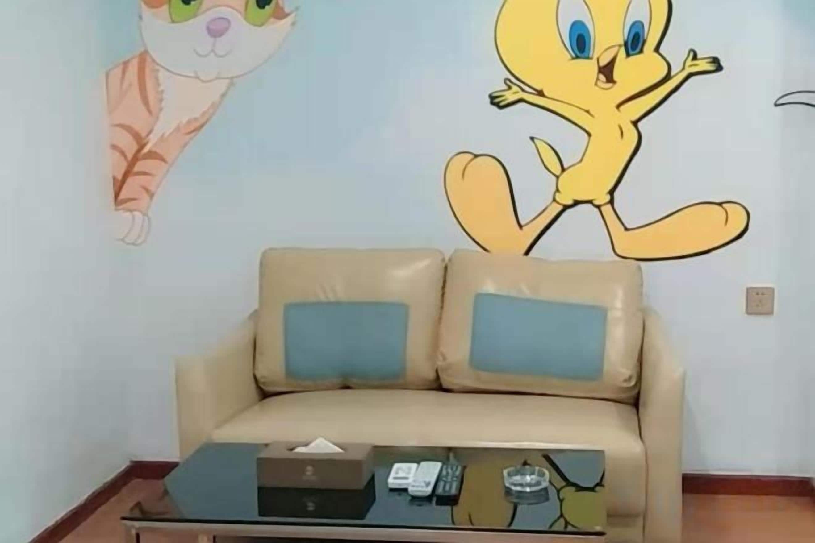 A beige sofa with two blue cushions, a glass coffee table with a remote control, and a wall with cartoon characters.