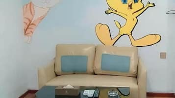 A beige sofa with two blue cushions, a glass coffee table with a remote control, and a wall with cartoon characters.