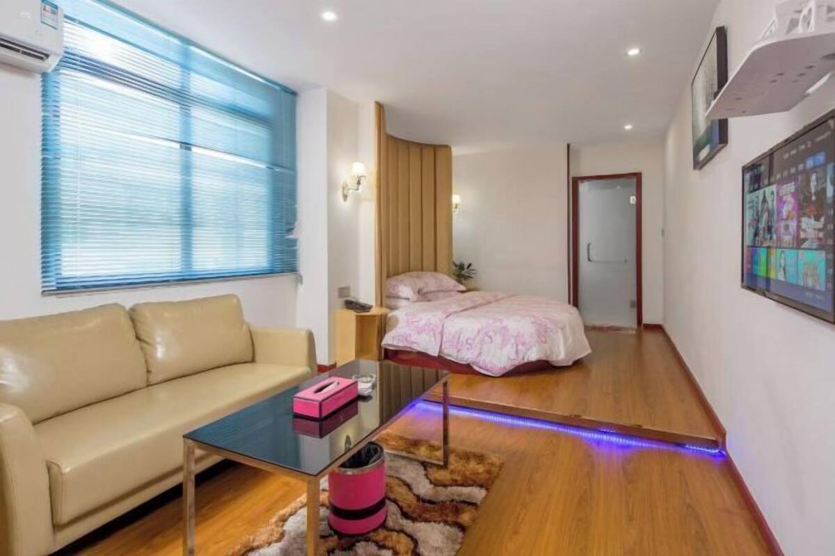 Une chambre d’hôtel moderne avec un canapé, un lit recouvert d’une courtepointe rose, un plancher en bois, une table basse en verre et un téléviseur fixé au mur.