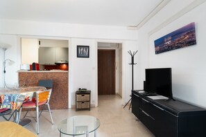 Ein Wohnzimmer mit Glastisch, Fernseher auf schwarzem Standfuß, Essbereich mit Tisch und Stühlen sowie Küchenzeile mit Arbeitsplatte und Geräten.
