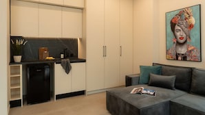 Une cuisine moderne avec un plan de travail noir, un lave-vaisselle encastré et un grand placard. On y trouve un canapé gris avec des coussins turquoise et un magazine posé dessus. Un tableau représentant une femme en robe colorée est accroché au mur.