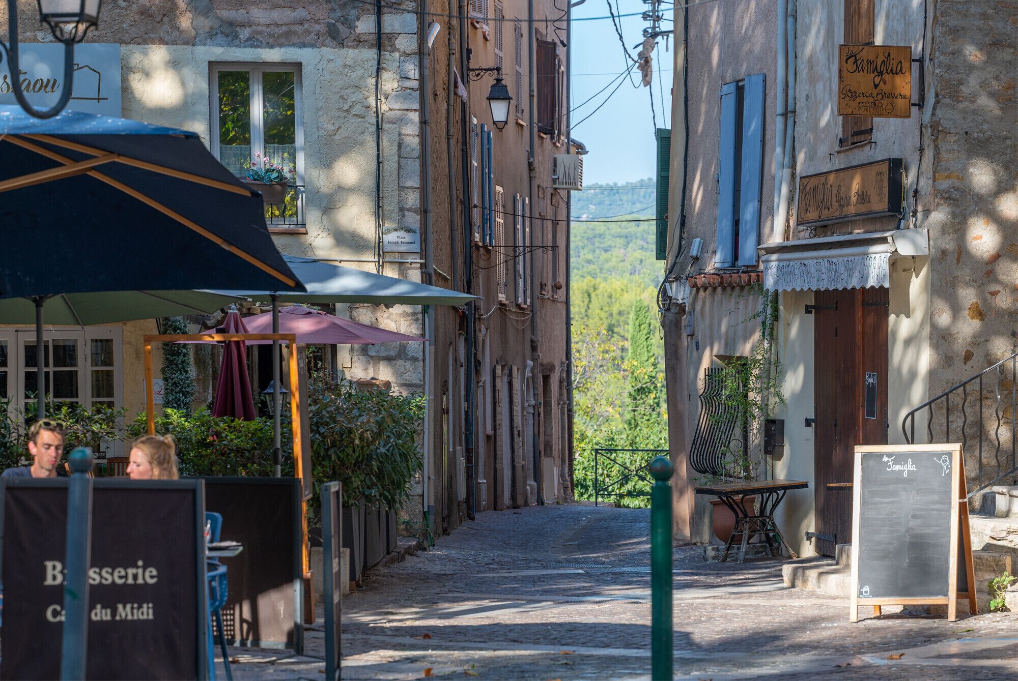 A narrow street with buildings, outdoor seating, and signs indicating local businesses.