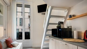 Eine kleine Küche mit einer Mikrowelle, einem Toaster und einem Holzbodenregal, ein Sofa mit Kissen und ein Fenster mit Blick auf ein Gebäude.