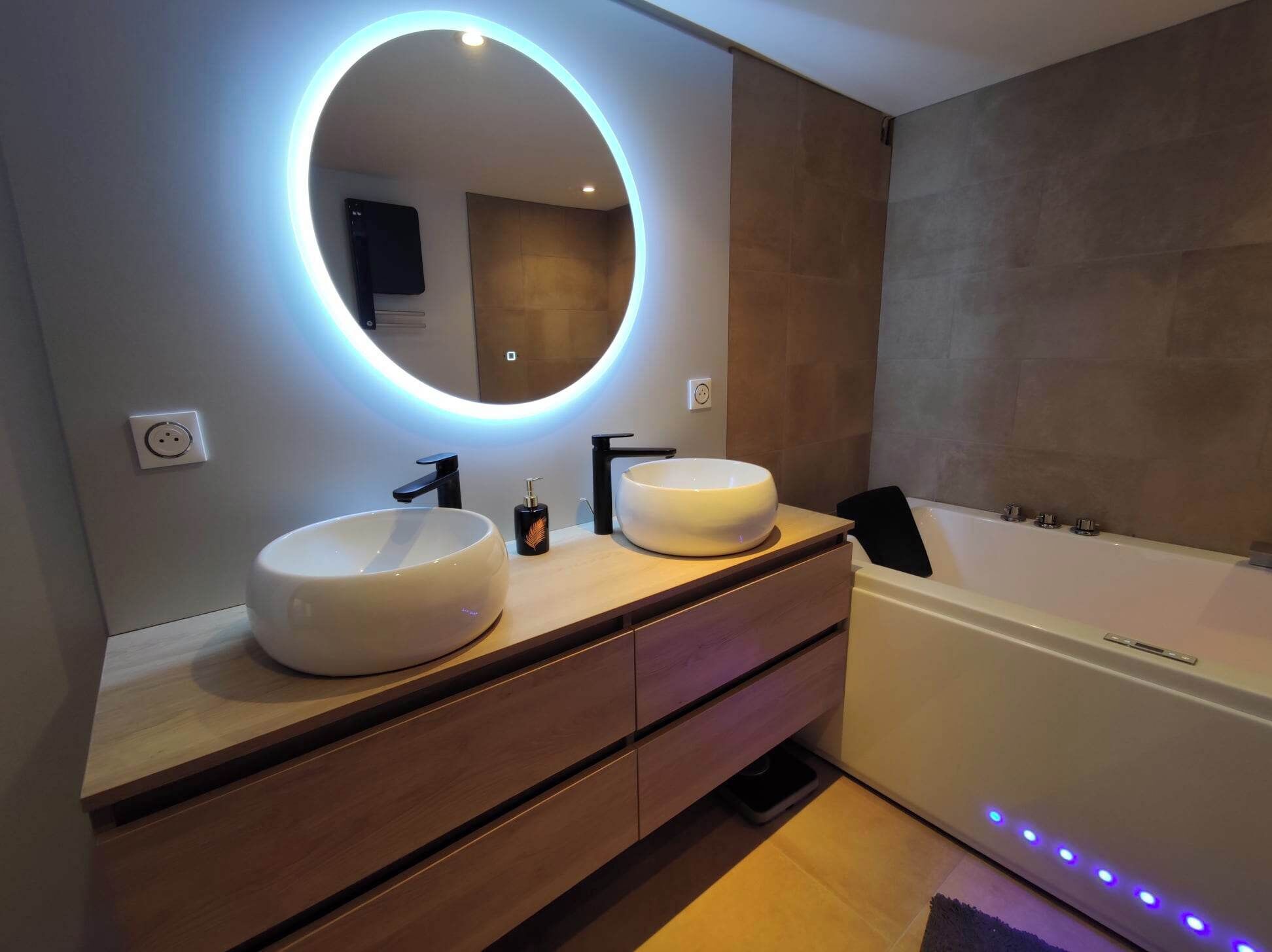 A modern bathroom with two sinks, a large mirror with underglow lighting, a bathtub, and a wooden vanity.
