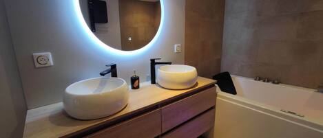 A modern bathroom with two sinks, a large mirror with underglow lighting, a bathtub, and a wooden vanity.