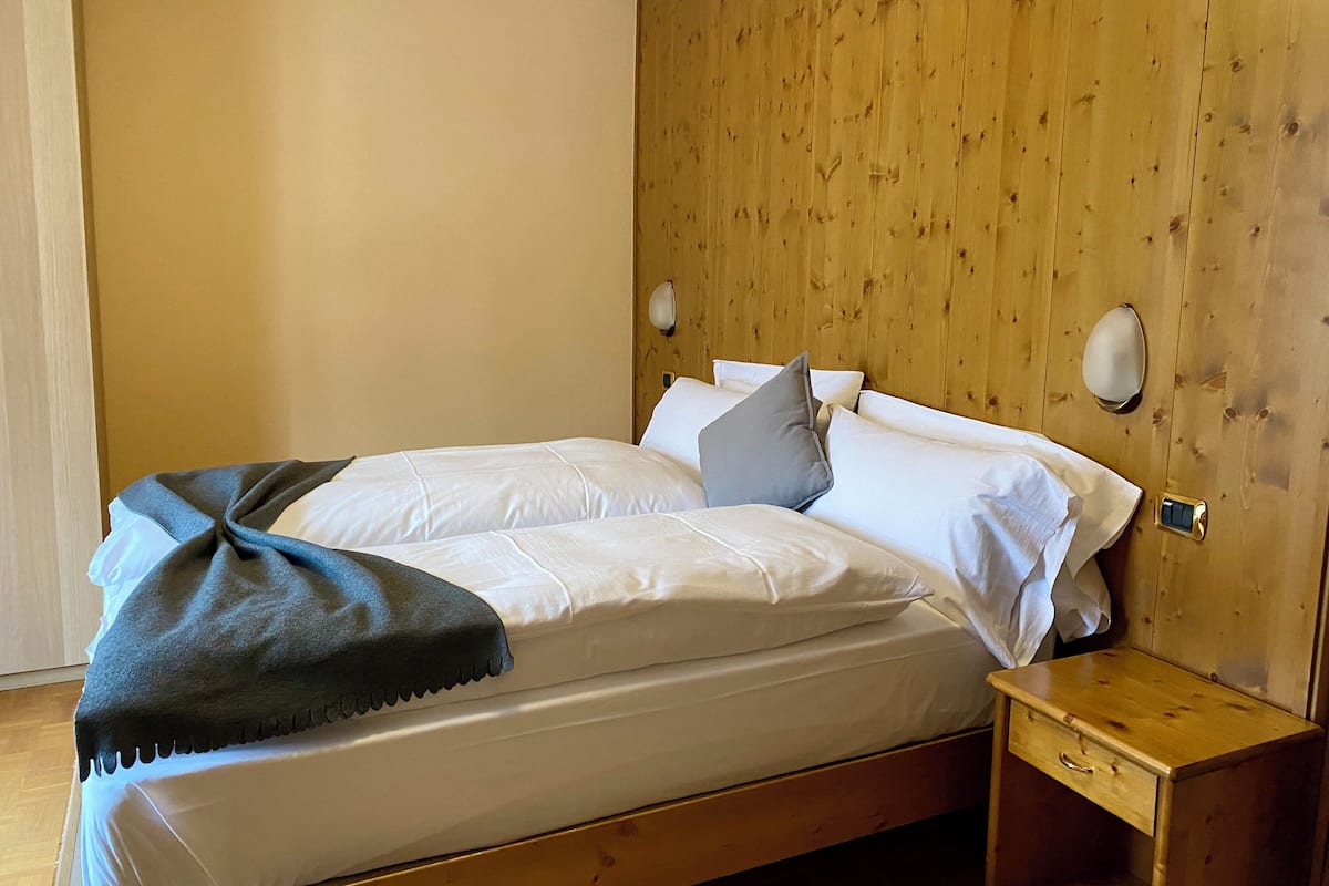 Una cama bien tendida con sábanas blancas y un edredón gris, una mesita de noche de madera con un cajón, y paneles de pared de madera.