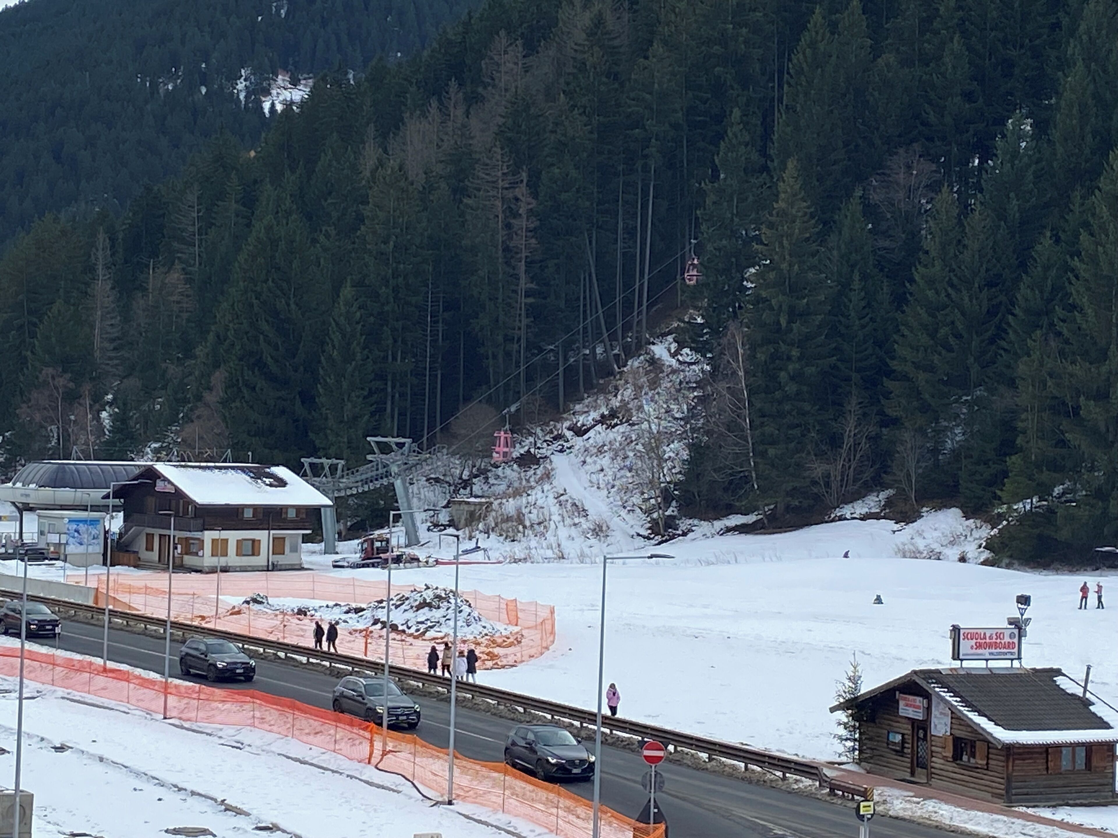 Paisaje nevado con un telesilla, una carretera con barreras naranjas y un pequeño edificio de madera con un letrero.