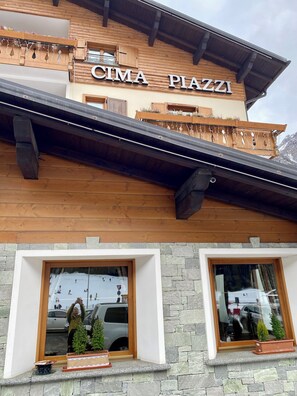 A building with a wooden facade and the name "CIMA PIAZZI" prominently displayed. There are two windows with stone frames, and potted plants are placed on the ledge below. - Hotel Cima Piazzi  (Valdidentro)