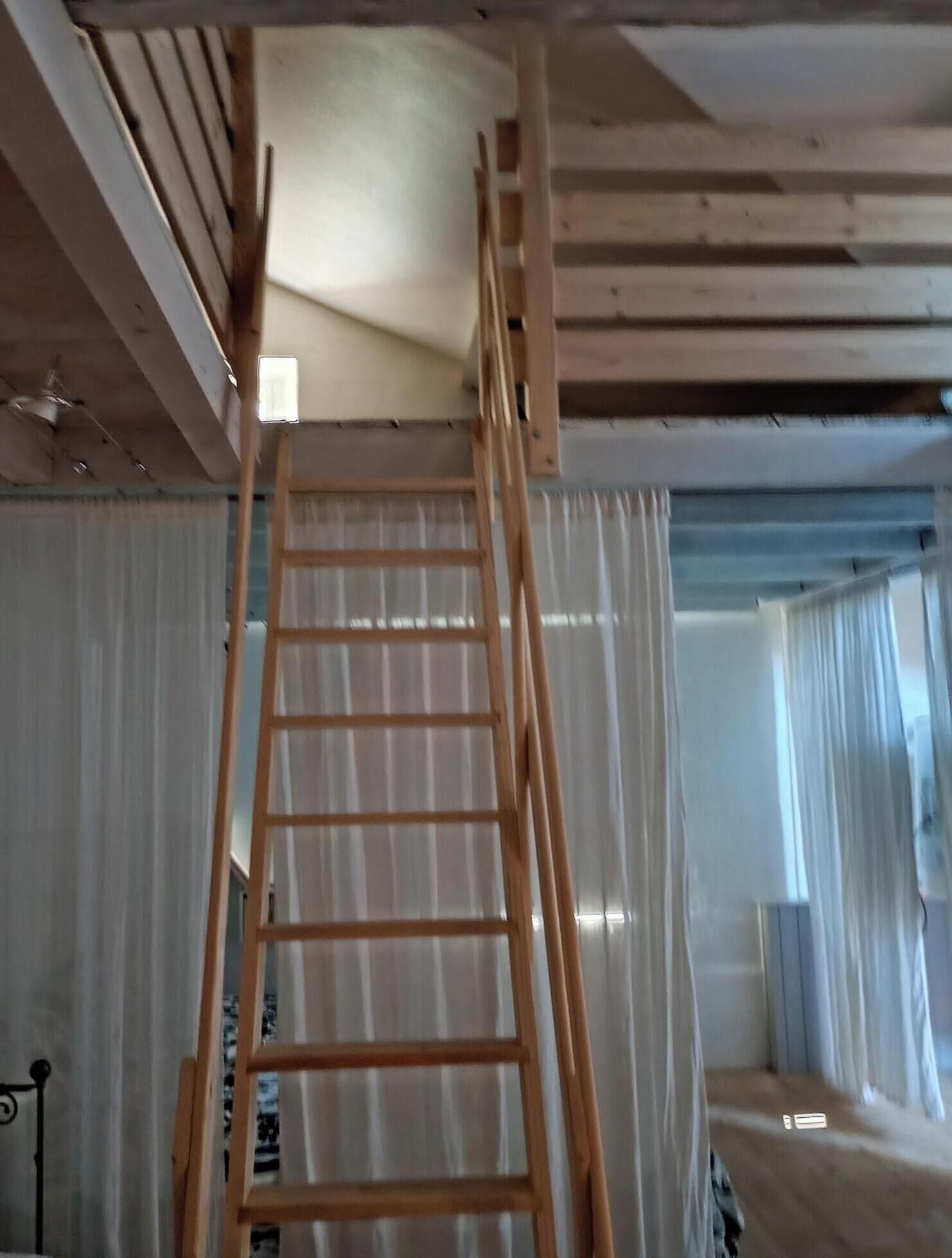 A wooden ladder in a room with a sloped ceiling, a window with curtains, and a metal railing.