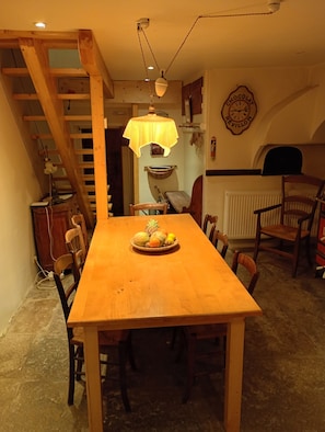 A wooden dining table with chairs, a fruit bowl, a hanging light fixture, a wooden staircase, and a wall clock.