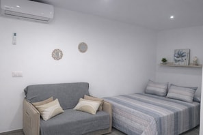 Ein Schlafzimmer mit grauem Sofa, einem Bett mit gestreifter Bettwäsche, einer Wandklimaanlage und einem kleinen Regal mit Dekoration.
