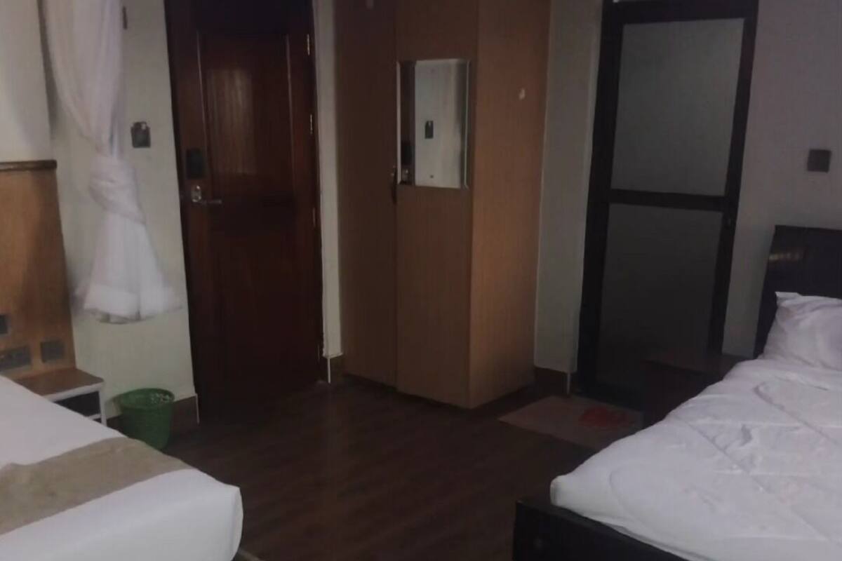 A hotel room with two beds, wooden flooring, a green trash bin, a wooden wardrobe, and a mirror on the wall.
