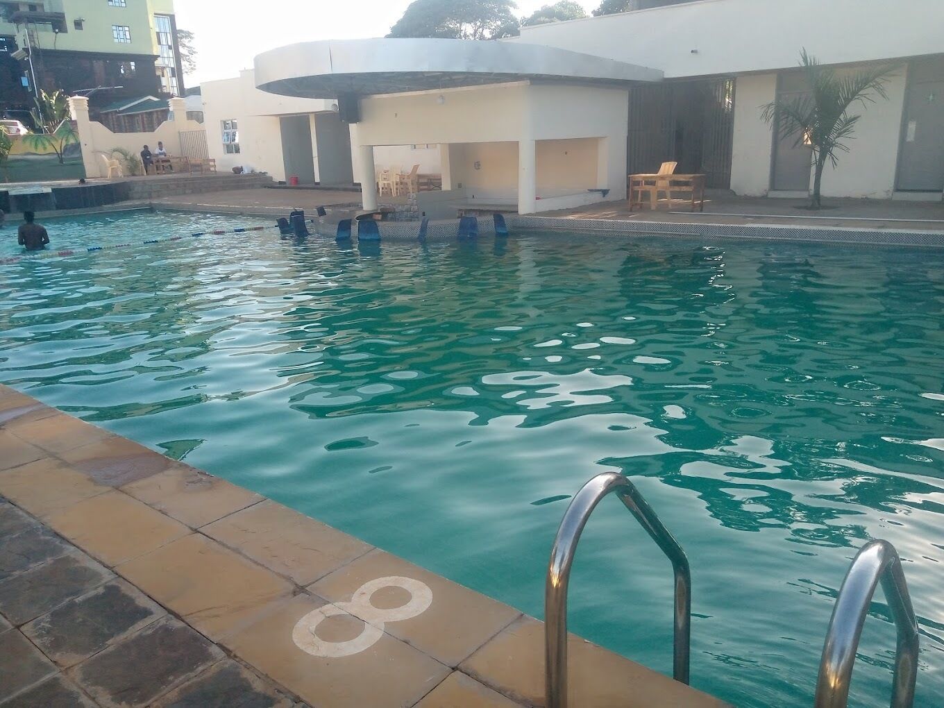 En swimmingpool med klart vand, omgivet af en flisebelagt terrasse og en bygning med udendørs siddepladser.
