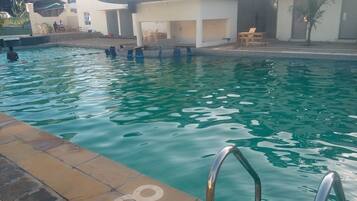 En swimmingpool med klart vand, omgivet af en flisebelagt terrasse og en bygning med udendørs siddepladser.