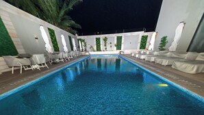 Una piscina con sillones de descanso y sombrillas, rodeada por un muro bajo con vegetación y paneles blancos.