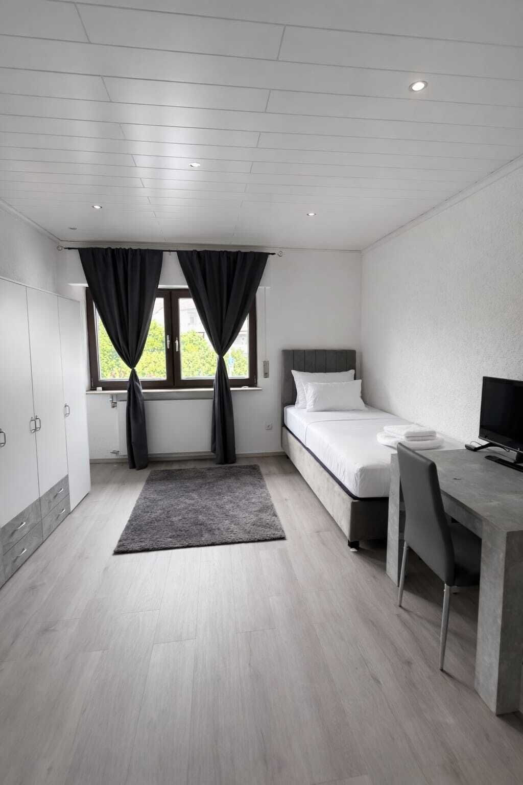 Un dormitorio moderno con cama, escritorio y silla, una ventana con cortinas y vista a espacios verdes.