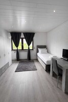 Un dormitorio moderno con cama, escritorio y silla, una ventana con cortinas y vista a espacios verdes.