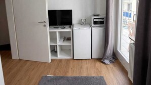 A compact room with a TV, a small fridge, a microwave, and a desk with a chair. There's a window with curtains and a view of buildings outside. - Frankfurt West Family Stay (Kelsterbach)