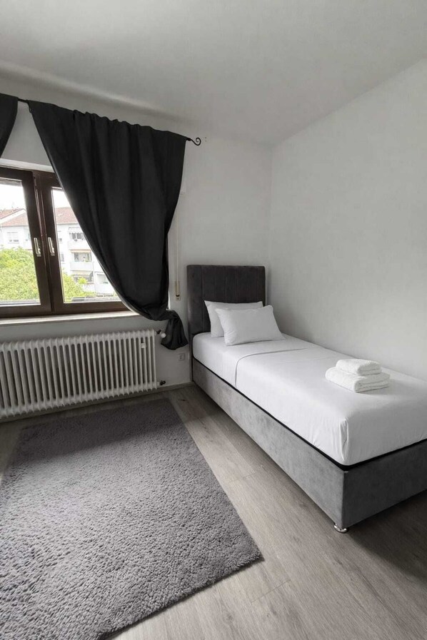 A small, modern bedroom with a single bed, a radiator, a grey rug, and a window with curtains. - Frankfurt West Family Stay (Kelsterbach)