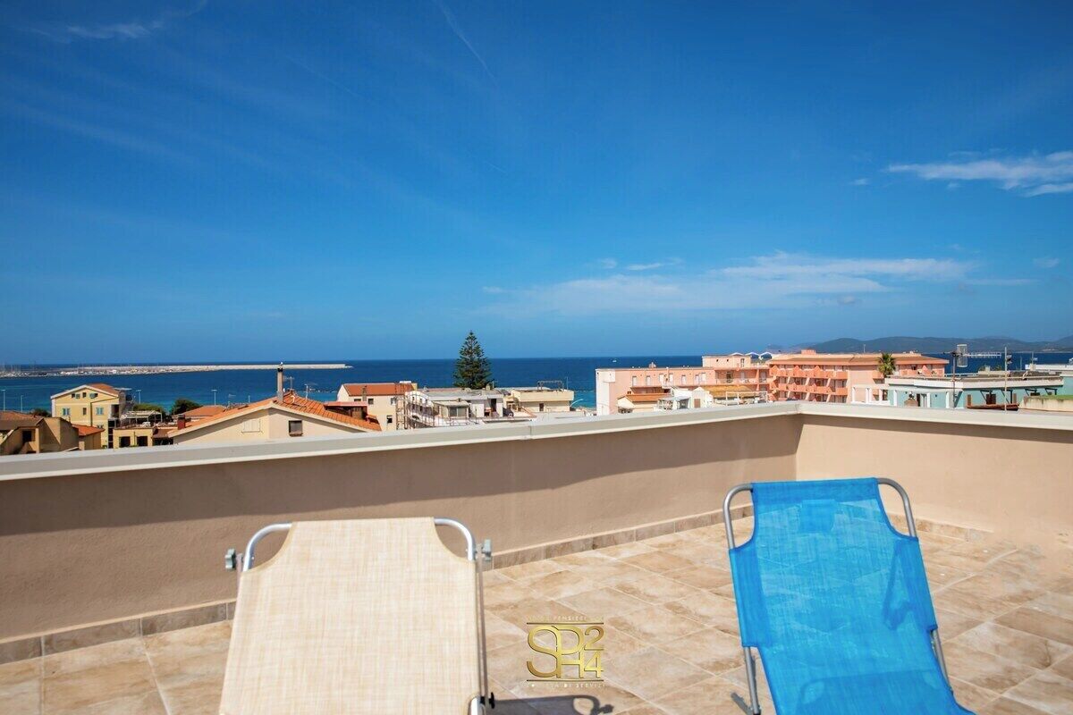 A rooftop terrace with two sun loungers, a clear view of the sea, and a row of buildings in the background.
