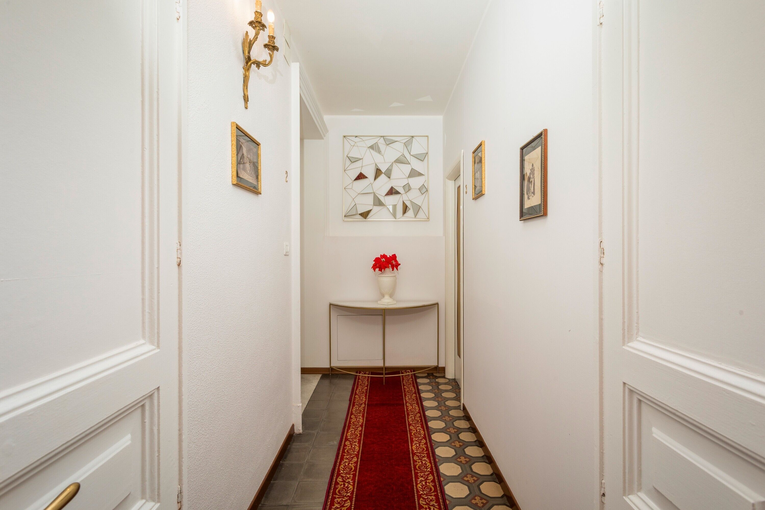 En hall med vita väggar, en röd matta, en geometrisk väggdekoration och ett vitt konsolbord med en vas med röda blommor.